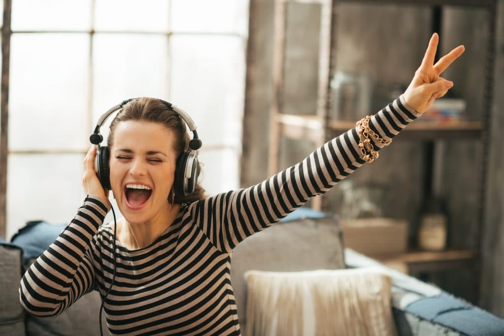 Moça jovem vestindo uma blusa listrada de preto e cinza com mangas longas. Ela está ouvindo a sua música faorita com fones de ouvido.
