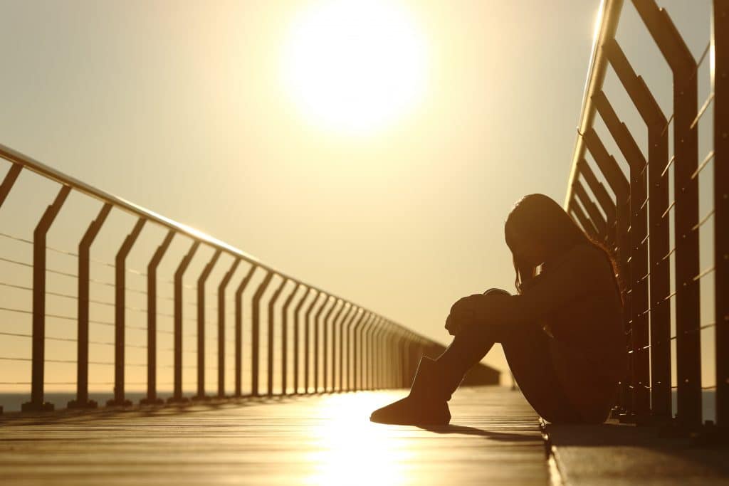 Imagem de uma mulher triste e cabisbaixa sentada sobre uma passarela. Ao fundo uma forte luz solar.