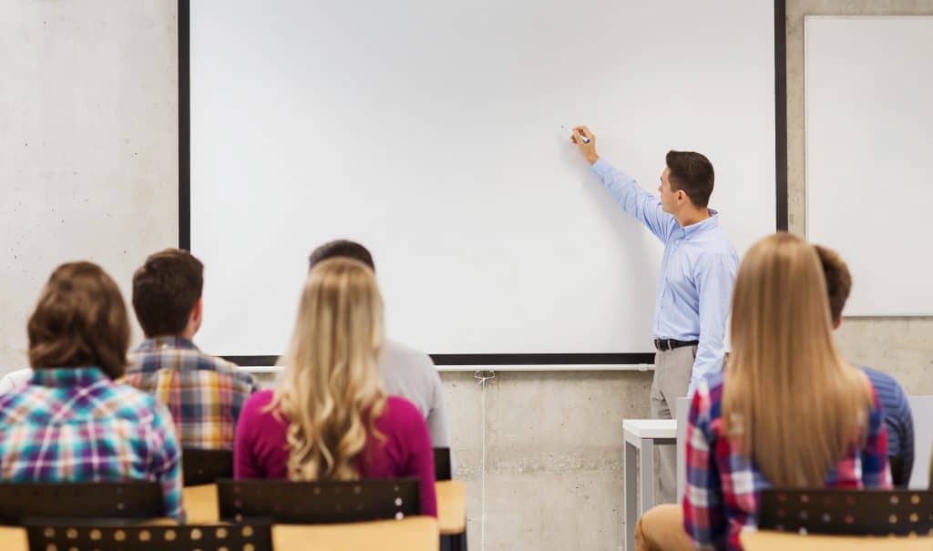 Vários alunos dentro de uma sala de aula. Todos estão prestando atenção na aula do professor que está escrevendo algo no quadro branco.