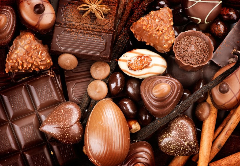 Vários tipos de chocolate em diversos formatos: em barra, em ovos de pásgoa, em bombons, em corações. Estão todos juntos dispostos sobre uma mesa decorada com canela em pau.