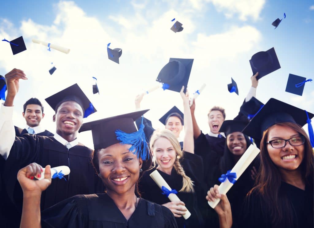 Vários alunos em ritmo de festa, pois estão se formando. Eles usam beca e estão jogando o capelo para cima. São negros, brancos e japoneses, segurando em suas mãos o diploma.