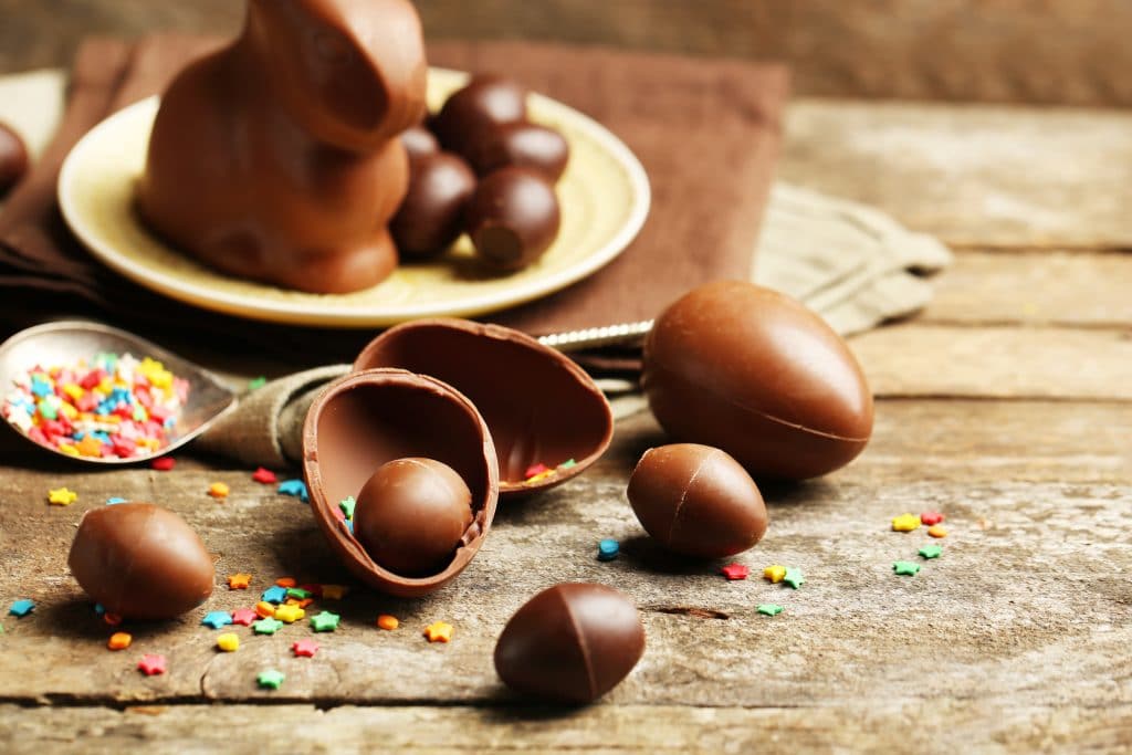 Ovos de páscoa de chocolate de vários tamos. Alguns estão inteiros, outros pela metade. Ao fundo um prato com alguns ovinhos e um coelho feito de chocolate.