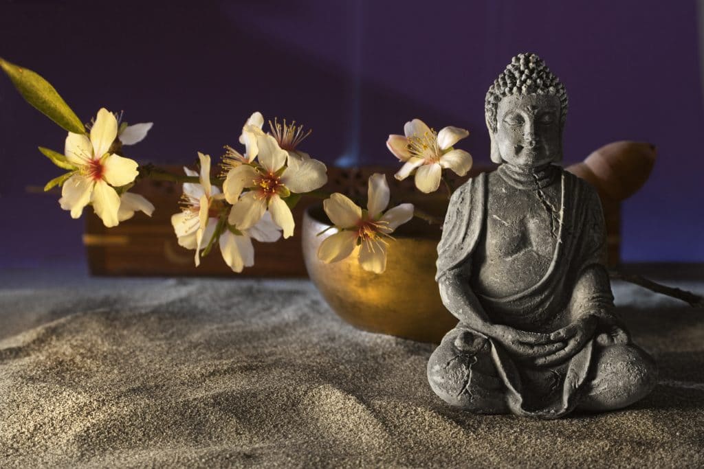 Imagem de um buda sentado. Ao lado flores e uma tigela tibetana dourada.
