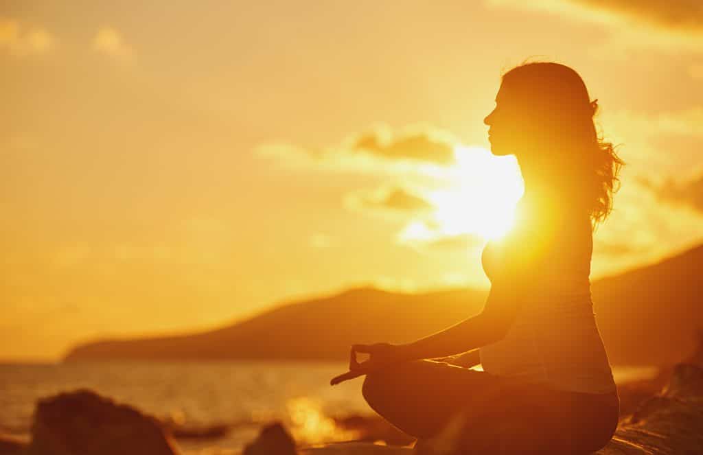 Mulher grávida praticando ioga, sentada em posição de lótus na praia e apreciando o pôr do sol.