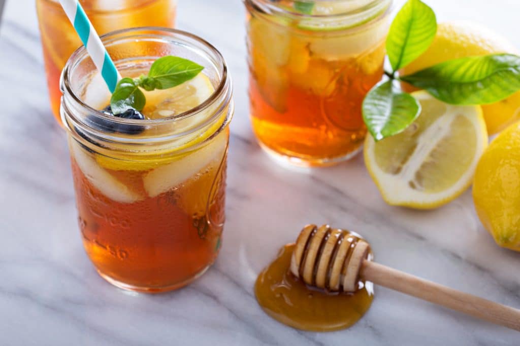 Suco de laranja com própolis e limão. Ele está sendo servido em canecas de vidro. Estão decorados com limão em pedaços e folha do limão. Ao lado temos uma colher de madeira contador de gotas de mel.
