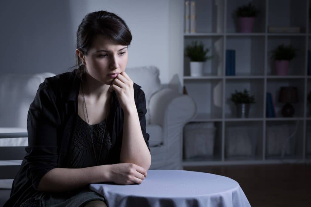 Mulher sentada. Ela está com os braços apoiados sobre uma mesa redonda com toalha branca. Ela está triste e deprimida.
