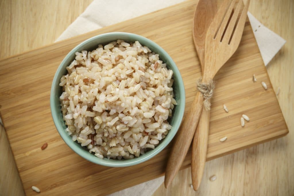 Arroz integral preparado e servido em um tigela azul. Ele está sobre uma mesa de madeira e ao lado um garfo e uma colher de madeira finalizam a decoração.
