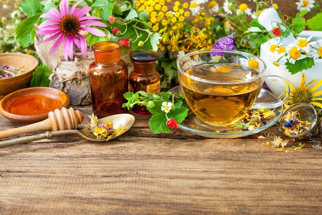 Chá de camomila dentro de uma xícata de vidro com pires. Ele está servido sobre uma mesa de madeira toda decorada com uma variedade de flores e mel.
