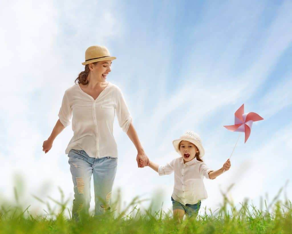 Mãe e criança feliz. Elas estão de mãos dadas caminhando em um campo bem verdinho. Elas estão vestidas iguais (chapéu de palha, camisa branca e jeans). A garotinha segura um catavento em uma das suas mãos.