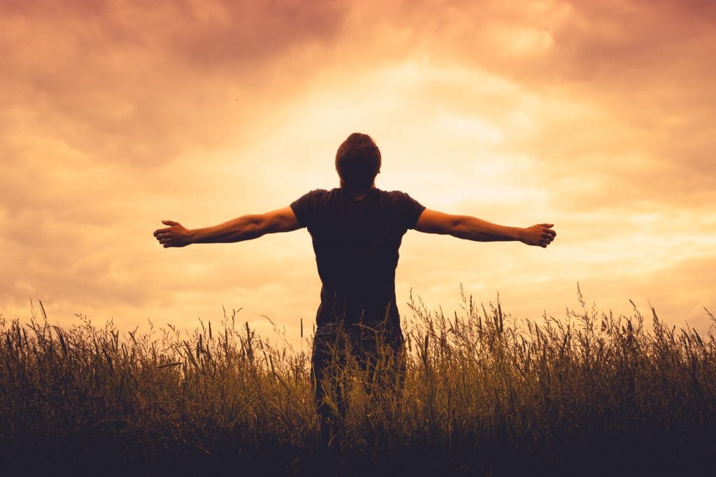 Homem de costas e de braços abertos fazendo uma oração de gratidão olhando para o sol se pondo.