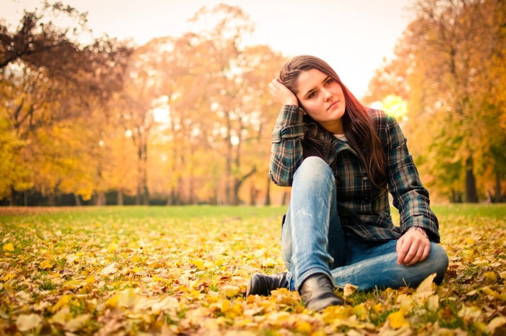 Mulher em estado depressivo. Ela está em um campo sentada sobre folhas caídas no chão. Ela usa calça jeans e camisa xadrez. 