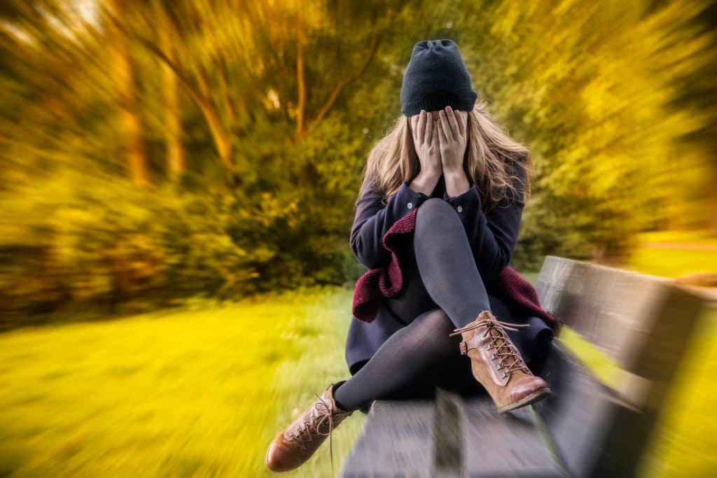 Imagem de uma mulher triste e com as duas mãos sobre o rosto. Ela está sentada em um banco no campo. Está frio. Ela usa roupas de inverno e uma touca de lã preta.