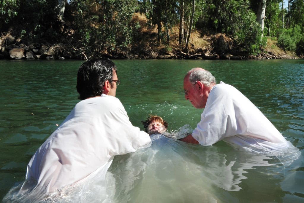 Dois homens com vestes branca reallizando o batismo de uma criança dentro de um rio cercado de uma mata verde.