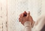 Mãos femininas entrelaçadas em frente a uma cortina branca de renda. Ela aparenta estar rezando.