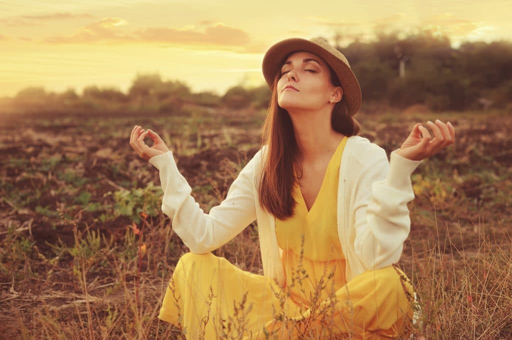 Mulher em posição de meditação. Ela está em um lindo campo cerrado. Ela veste um vestido amarelo, casaco de linha branco e um chapéu de palha.
