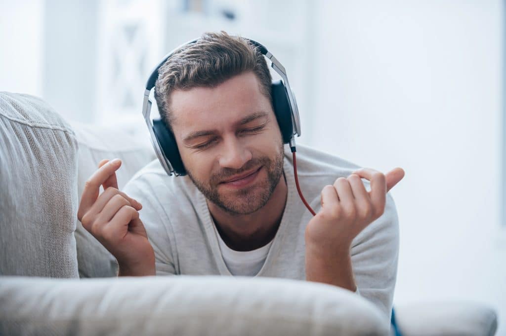 Jovem alegre usando seus fones de ouvido e apreciando a sua música favorita. Ele está alegre deitado de bruço no sofá de sua sala.