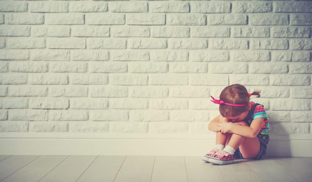 Criança menina, sentada no chão. Ela está chorando. Ao fundo uma parede de tijolinho branca.