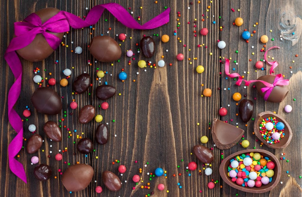 Ovos de Páscoa de chocolate de diversos tamanhos, inteiros e cortados ao meio, recheados com confetes. Eles estão sobre fundo de madeira rústico.
