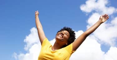 Mulher em pé sorrindo com braços para cima e céu azul com nuvens ao fundo