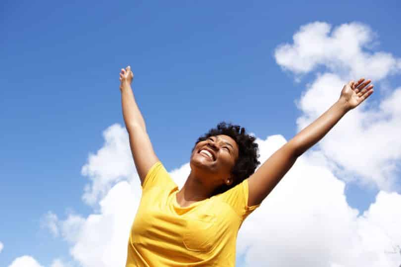 Mulher em pé sorrindo com braços para cima e céu azul com nuvens ao fundo