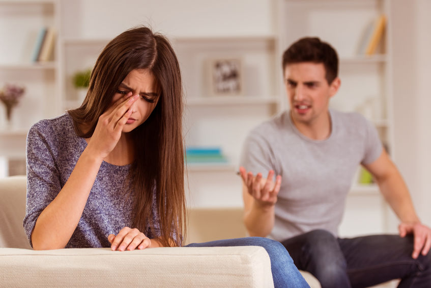 Homem com cara de bravo e  mão levantada falando com mulher, que está de costas para ele, e coloca a mão no rosto para enxugar suas lagrimas.