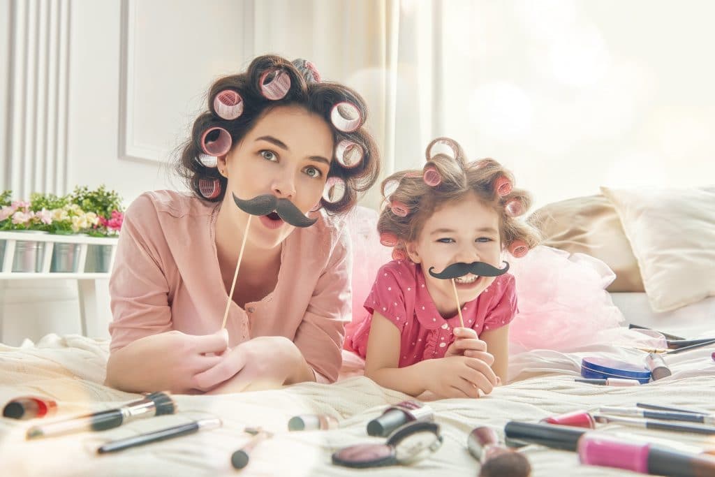 Mãe e filha deitadas sobre a cama. Elas estão usando bobs nos cabelos e segurando uma máscara de bigode. A cama está repleta de itens de maquiagem. É uma imagem alegre e descontraída onde ambas celebram o dia das mães.
