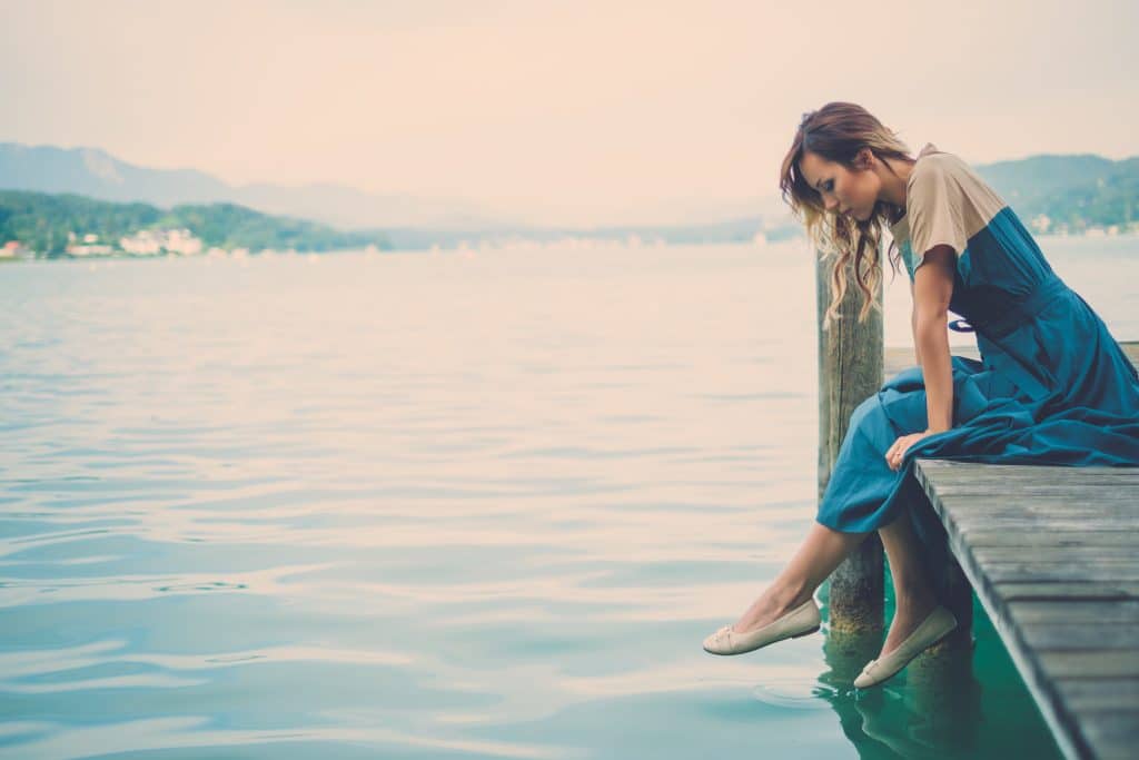 Mulher sentada na ponte e olhando para a água do lago. Ela está usando um vestido azul longo com mangas bege e uma sapatilha bege.
