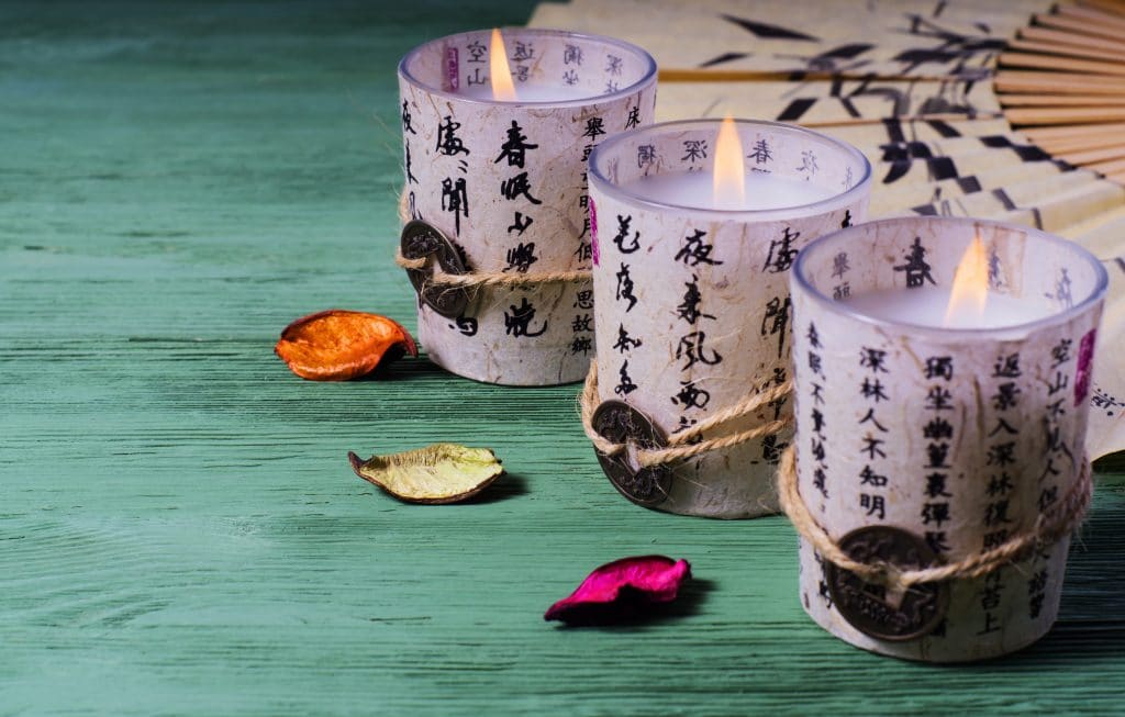 Velas em copos decorados com letras chinesas. Os copos estão sobre uma mesa de madeira pintada na cor verde. Ao lado um leque decora os mesmos.