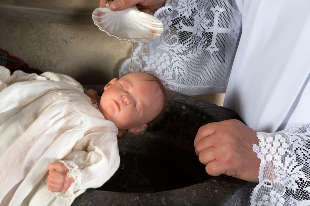 Criança sendo batizada na pia da igreja pelo padre. Ambos estão vestindo uma túnica branca. O padre usa em sua túnica uma barrado em renda.
