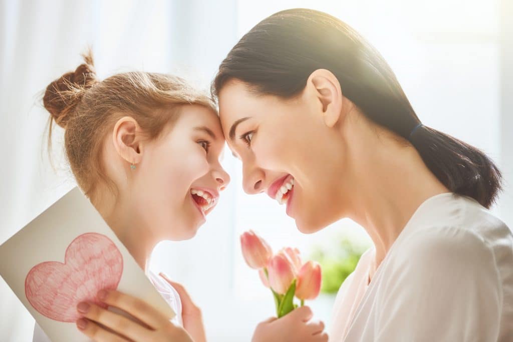 Mãe e filha se abrando e olhando uma para outra com um gesto de muito amor e carinho. A criança oferece um cartão com um coração pintado e um buquê de tulipas rosas.
