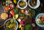 Mesa de madeira com muitas comidas vegetarianas como salada com picles, sanduíche com abacate, macarrão com grão de bico e quinoa.