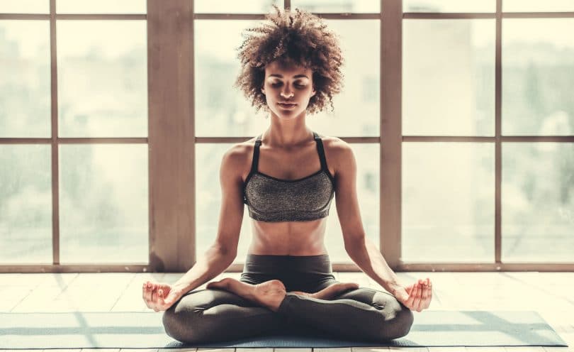 Mulher sentada em posição de meditação com olhos fechados e janelas iluminadas ao fundo