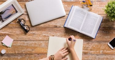 Pessoa estudando em uma mesa com um livro, um caderno, um notebook, quadros e plantas.