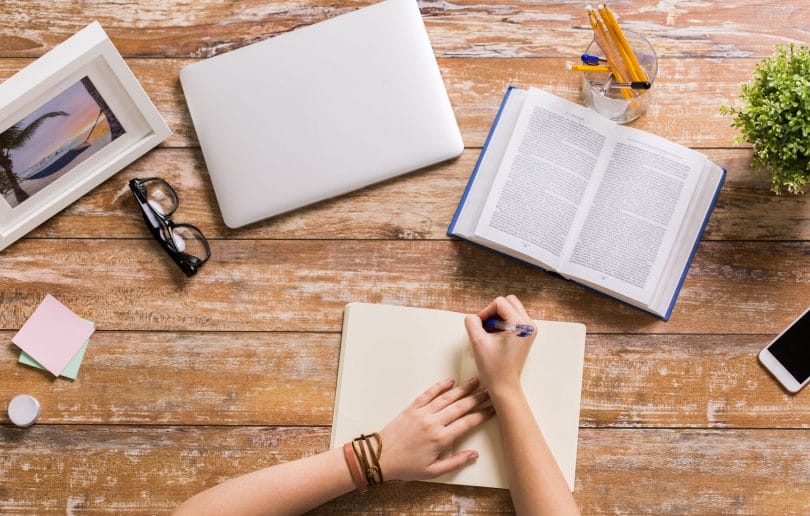 Pessoa estudando em uma mesa com um livro, um caderno, um notebook, quadros e plantas.