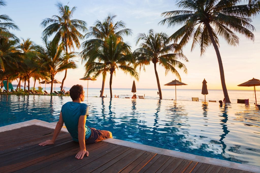 Homem vestindo bermuda marrom e camiseta azul. Ele está sentado no piso de madeira em frente da piscina. Ele está relaxando e observando o pôr do sol. 