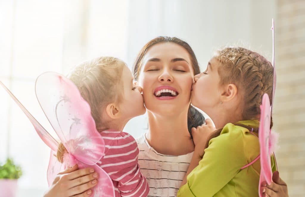 Mãe feliz comemorando o seu dia. Ela está sendo beijada pelas suas duas filhas que estão no seu colo. As crianças usam asas de borboleta na cor rosa.
