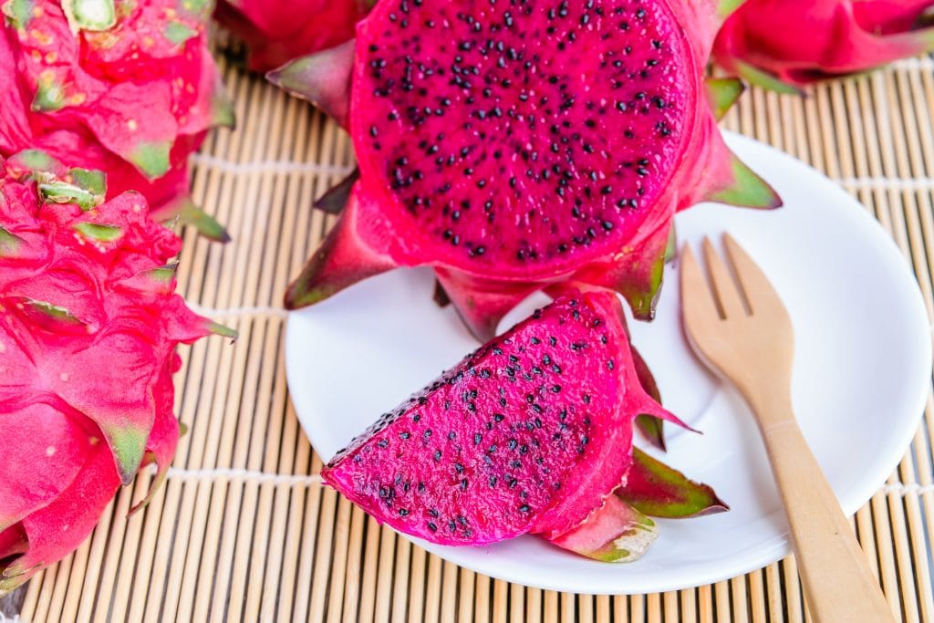 Fruta pitaya corta ao meio e servida sobre um prato de porcelana branco. Ao lado um garfo de madeira. O prato está sobre um suporte feito de madeira bege. Ao lado do prato também temos a fruta inteira.
