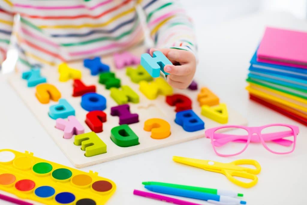 Mãos de uma criança montando um quebra-cabeça de letras coloridas. Sobre a mesa branca estão canetinhas, cadernetas, tesoura, óculos de grau e tinta guache. Tudo com muita cor.
