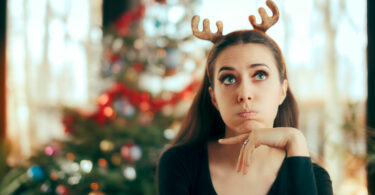 Mulher com arquinho de renas com expressão irritada e árvore de natal ao fundo