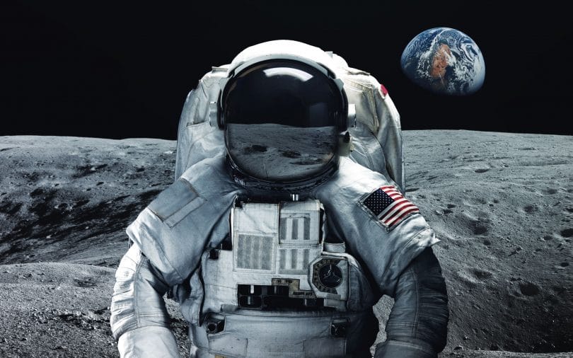 Astronauta com uniforme e bandeira dos Estados Unidos na Lua. Ao fundo é possível ver a Terra.