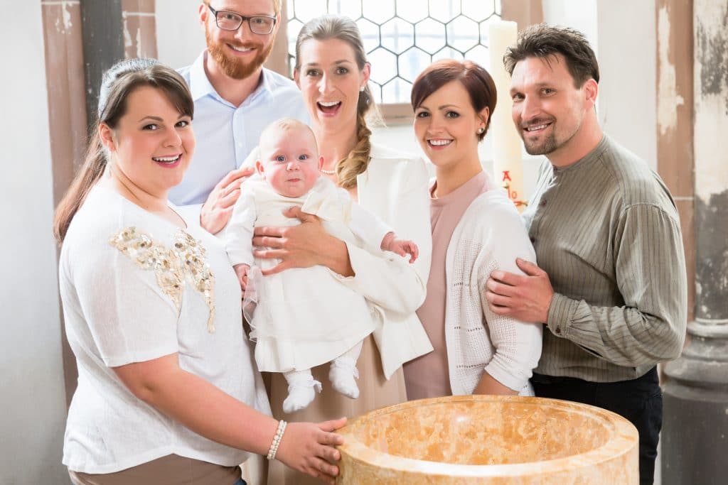 Família reunida para batizado de uma criança. Mãe segura o babê em seu colo e ele veste uma bata branca.