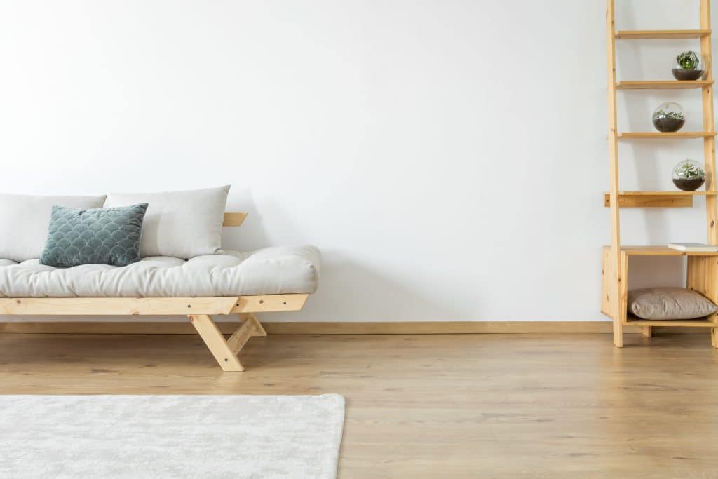 Escada de madeira clara. Ela serve de aparador de vasos de plantas. Ela está disponível em uma sala branca com piso de madeira clara. Ao lado da escada um sofá também de madeira. Ele tem algumas almofadas brancas e uma azul ao centro.