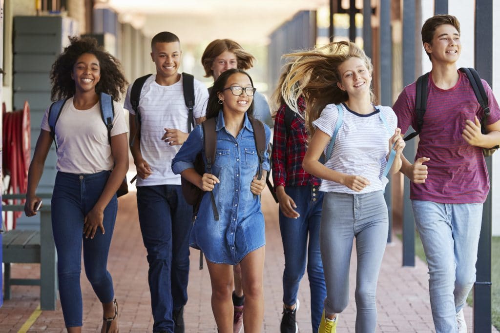 Grupo formado por jovens homens e mulheres de várias etnias. Eles estão com mochilas nas costas, sorrindo e andando no corredor da escola.