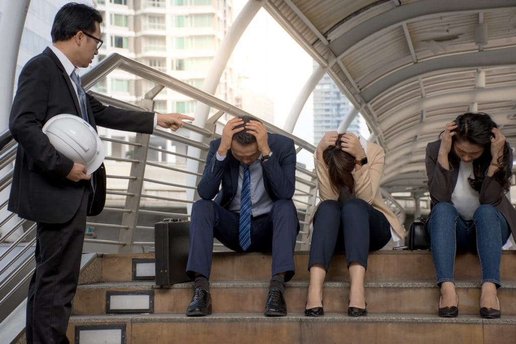 Homem de pé segurando um capacete e apontando o dedo para três pessoas sentadas, sendo duas mulheres e um homem. Os três estão de cabeça baixa com as mãos sobre ela. Eles estão sendo desprezados pelo homem que está de pé.