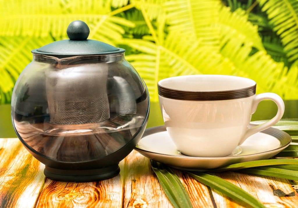 Chá de cardo-mariano servido em uma xícara e um pires de porcelana branco com uma faixa verde. Ao lado um bule de chá. As peças estão sobre uma mesa de madeira decorada com uma folhagem.