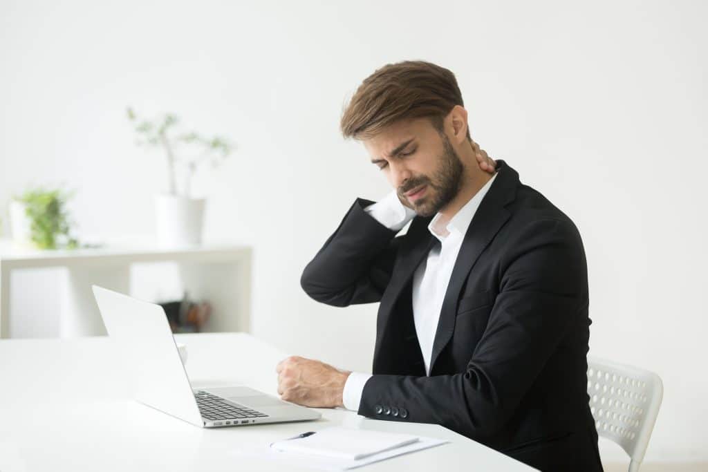 Homem de terno em uma sala do seu escritório Ele está de frente para o notebook. Aparentemente está com dores na nuca, pois uma de suas mãos está sobre ela.