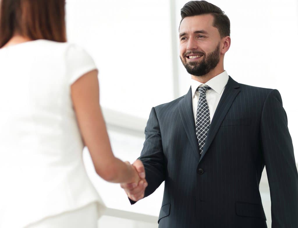 Homem de barba vestindo um terno cinza, camisa branca e gravata preta e branca cumprimentando uma mulher de cabelo curto, vestindo um conjunto de saia e blusa branca. Ambos estão felizes e gratos pelo investimento financeiro.