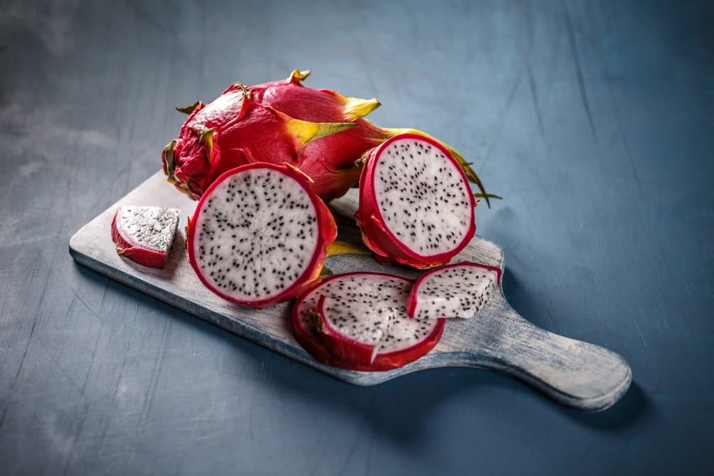 Várias pitayas sobre uma tábua de cortar carnes. Uma das frutas está inteira e a outra cortada ao meio e também fatiada.