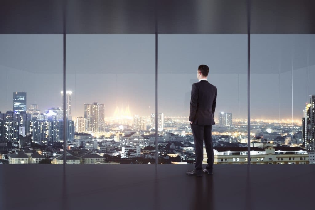 Homem jovem vestindo um terno preto. Ele está dentro de um escritório moderno com grandes janelas de vidro. Ele está olhando pela janela com uma vista da cidade à noite.