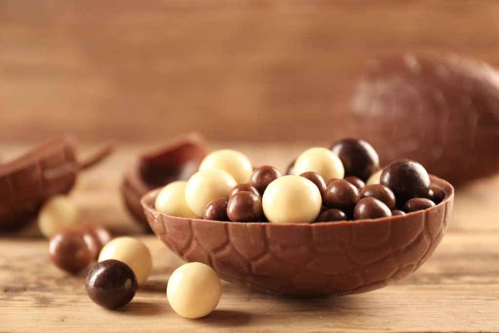 Ovo de Páscoa cortado ao meio e recheado com vários bombons de chocolate branco e ao leite. O ovo de páscoa está disposto em um fundo de madeira.